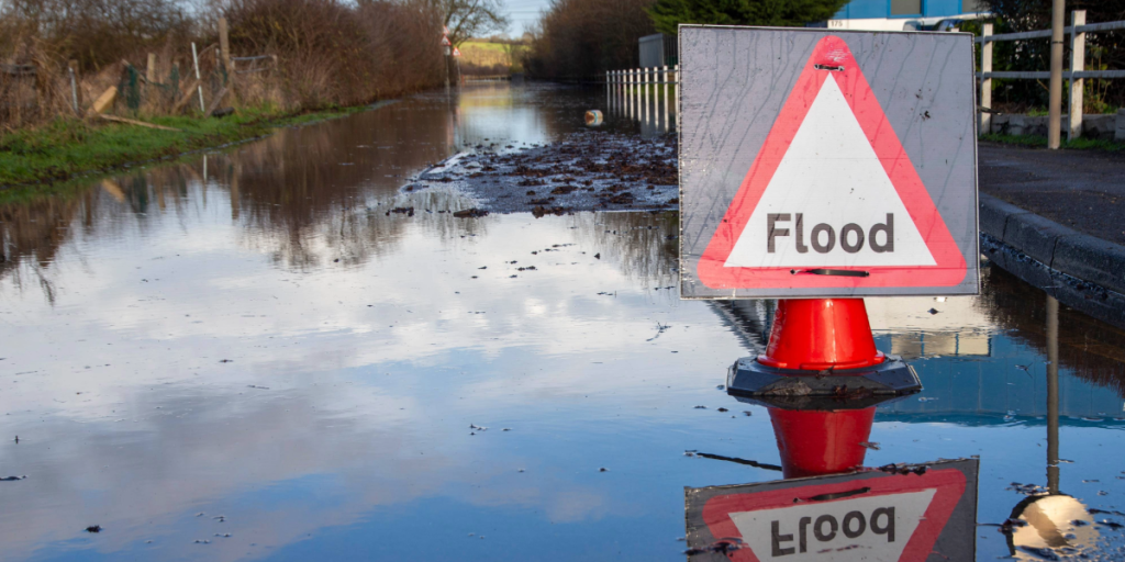 The image shows a road with surface water on and a sign which says 'flood'